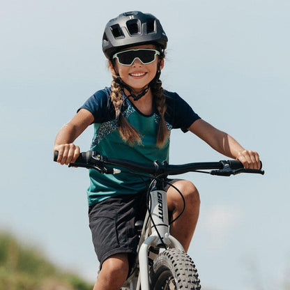 A young girl wearing a bike helmet, Julbo-usa Fury Mini Kids sunglasses, and a sports outfit rides her bicycle on a sunny outdoor pump track.