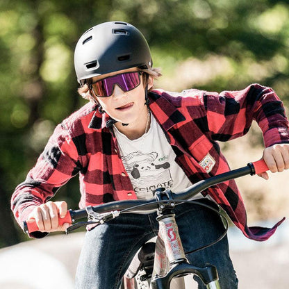A person wearing a helmet and Julbo-usa Fury S Kids sport sunglasses rides a bicycle outdoors in daylight, dressed in a red plaid shirt.