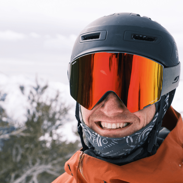 A person wearing a black ski helmet, reflective ski goggles, and a patterned neck gaiter smiles outdoors with snow and trees in the background.