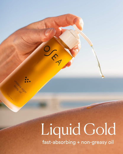 Hand applying OSEA liquid gold body oil with a blurred beach background